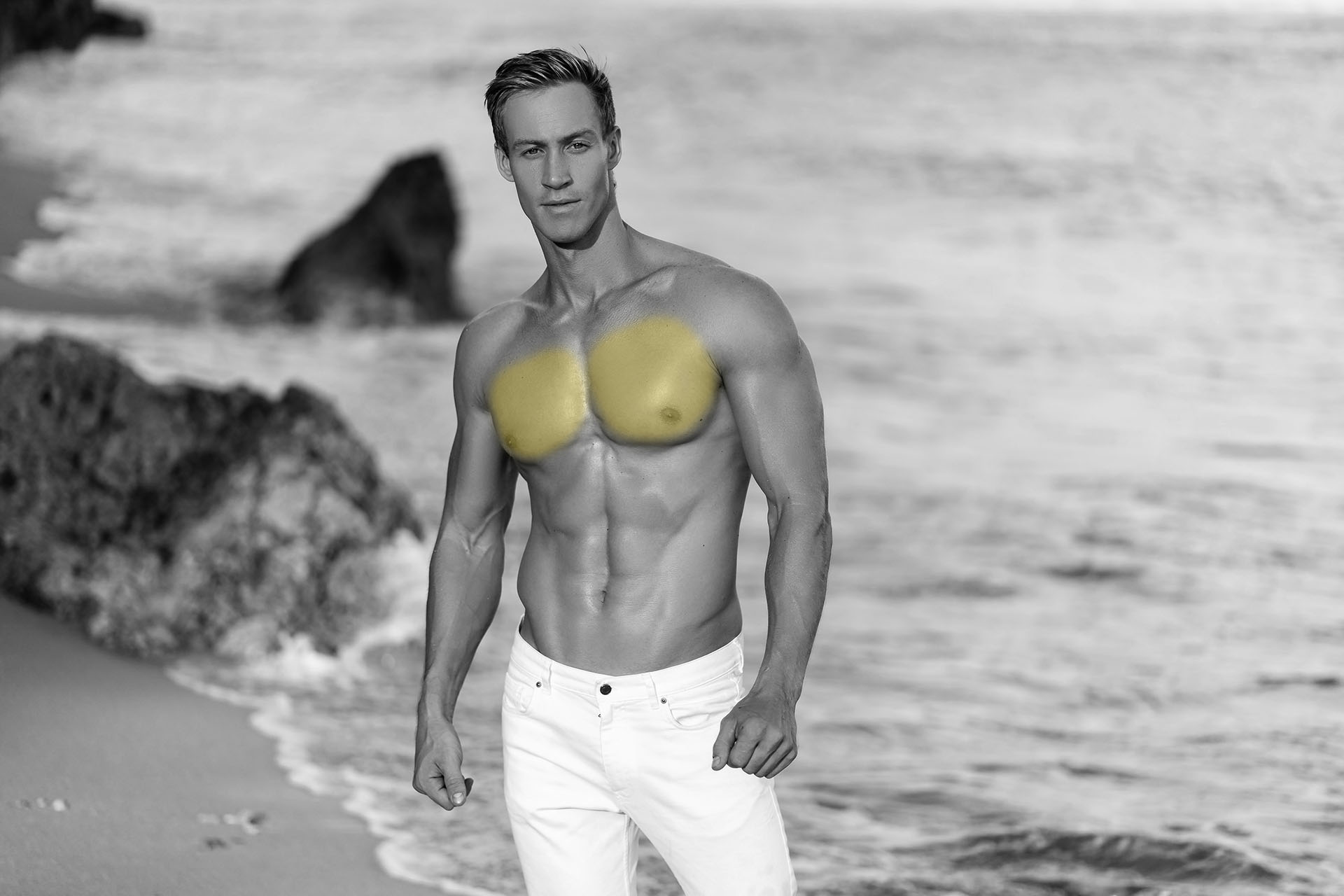 Sexy muscular guy in white pants and shirtless posing on tropical sandy beach, ocean waves at background